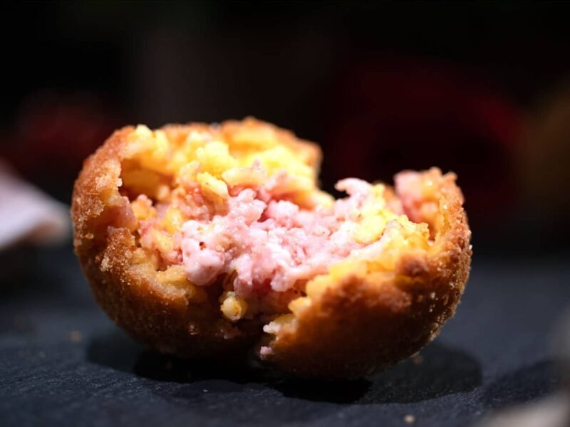 Arancino al prosciutto aperto a metà con ripieno di prosciutto ben visibile e poggiato su un supporto in ardesia