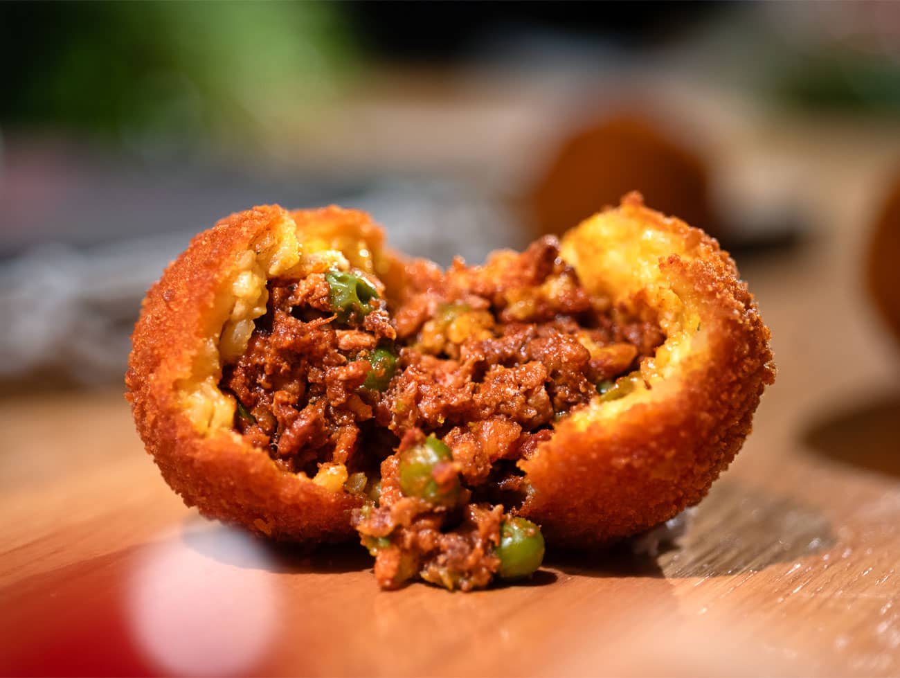 Immagine di un arancino classico con ripieno di ragù e piselli aperto a metà con ripieno ben visibile e poggiato su un tavolo