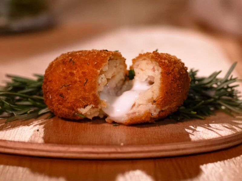 Foto di una crocchetta di patate alla brace poggiata su un tagliere di legno tondo e apeta a metà. Dietro alla crocchetta c'è un ramo di rosmarino per decorazione ed è visibile il ripieno.