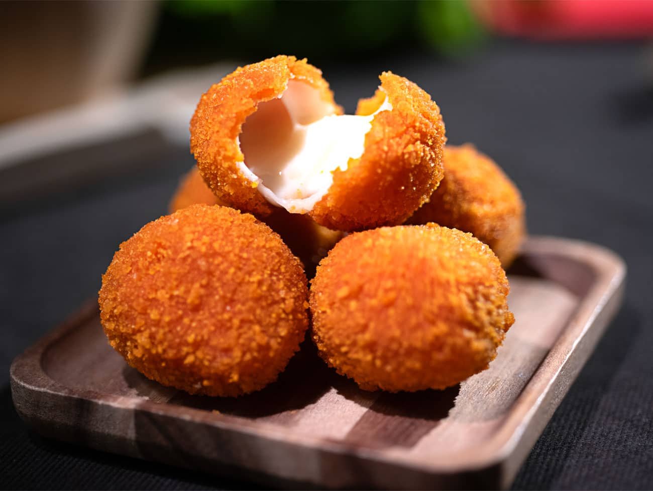 Foto di quattro mozzarelline fritte poggiate su un piattino di legno quadrato con angoli arrotondati. La mozzarellina che sta in alto è aperta a metà e svela la mozzarella fusa