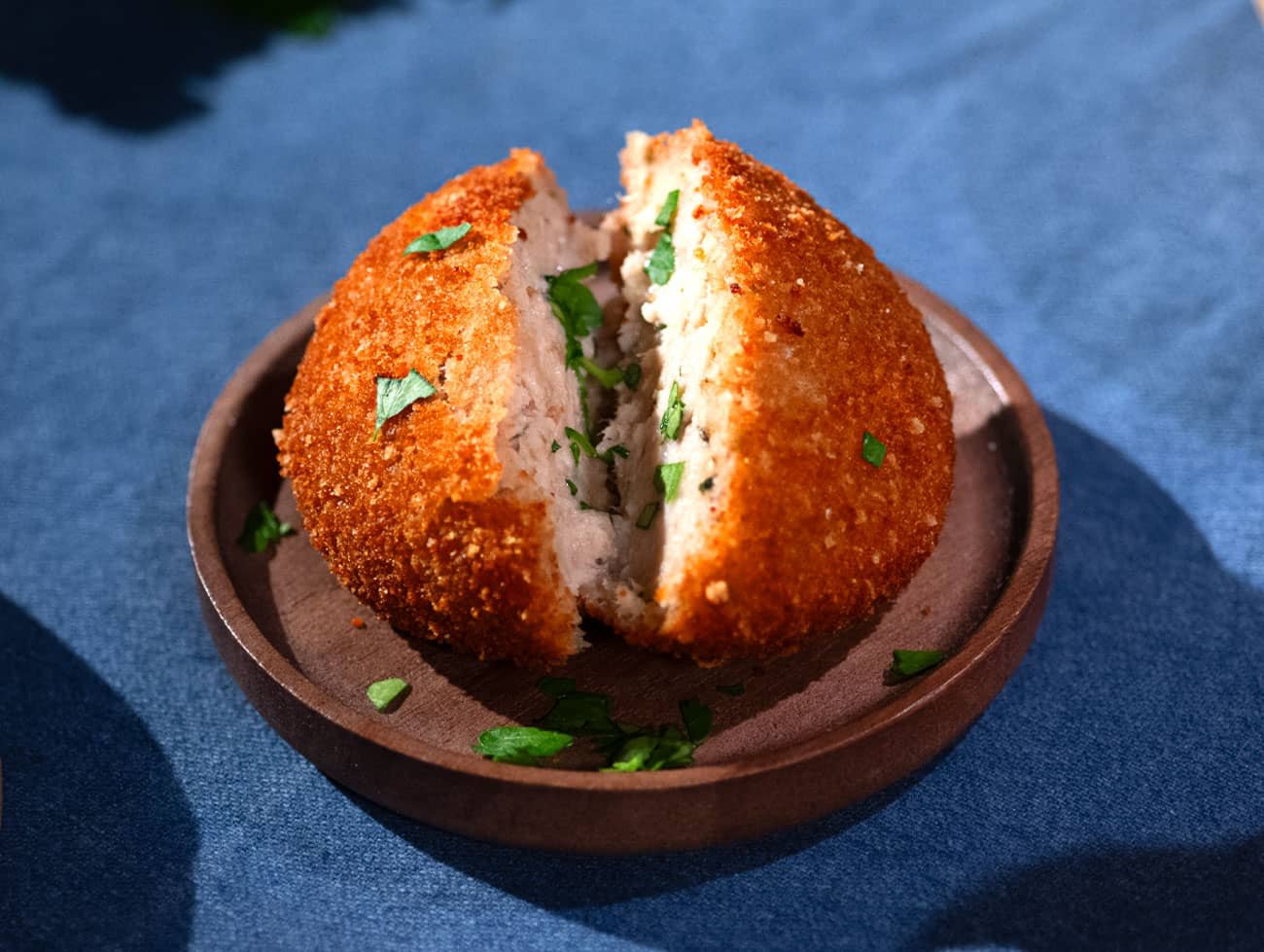 Foto di una polpetta di pollo aperta a metà poggiata su un piattino di legno, poggiato a sua volta su una tovaglia blu