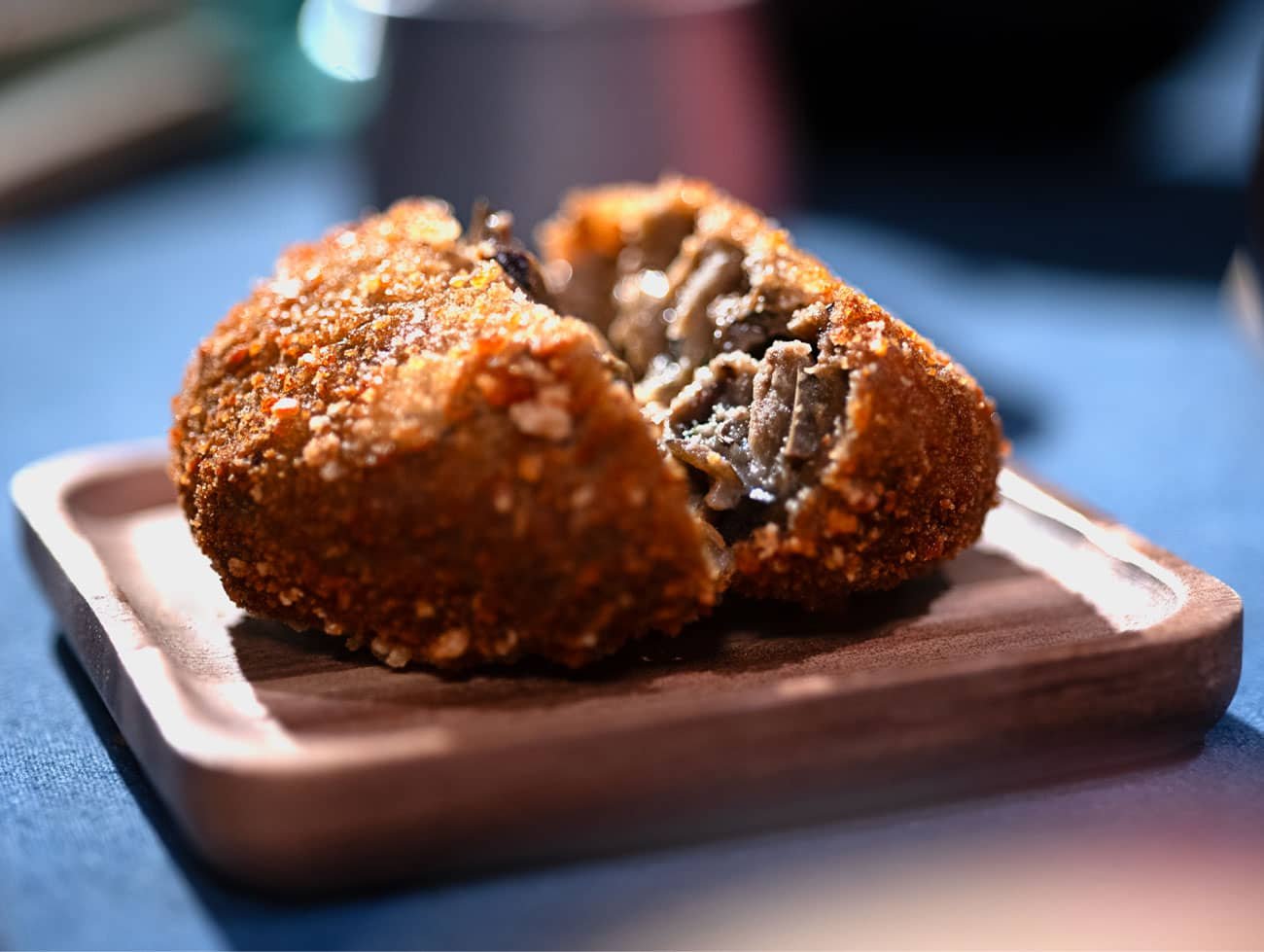 Foto di una polpetta di melanzane panata e tagliata a metà con il ripieno delle melanzane ben visibile. La polpetta è poggiata su un piattino quadrato di legno con angoli smussati
