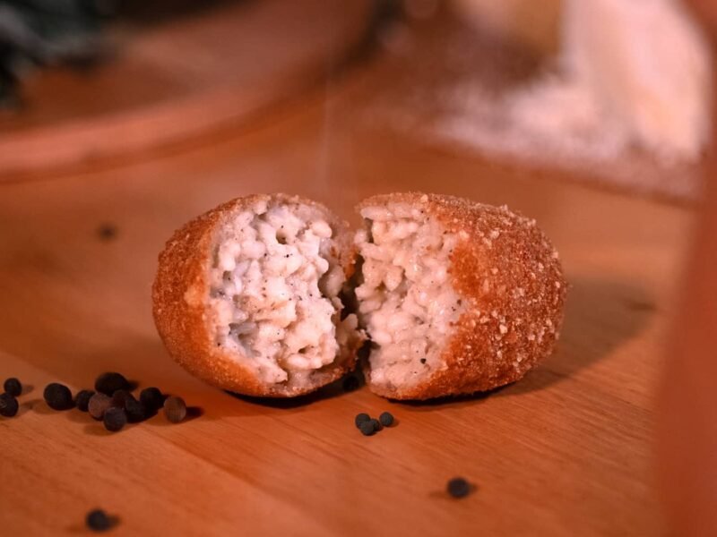 Supplì cacio e pepe aperto, poggiato su un tavolo con accanto dei grani di pepe come decorazione