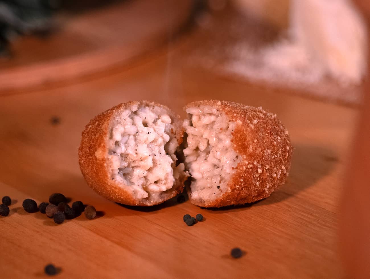 Supplì cacio e pepe aperto, poggiato su un tavolo con accanto dei grani di pepe come decorazione
