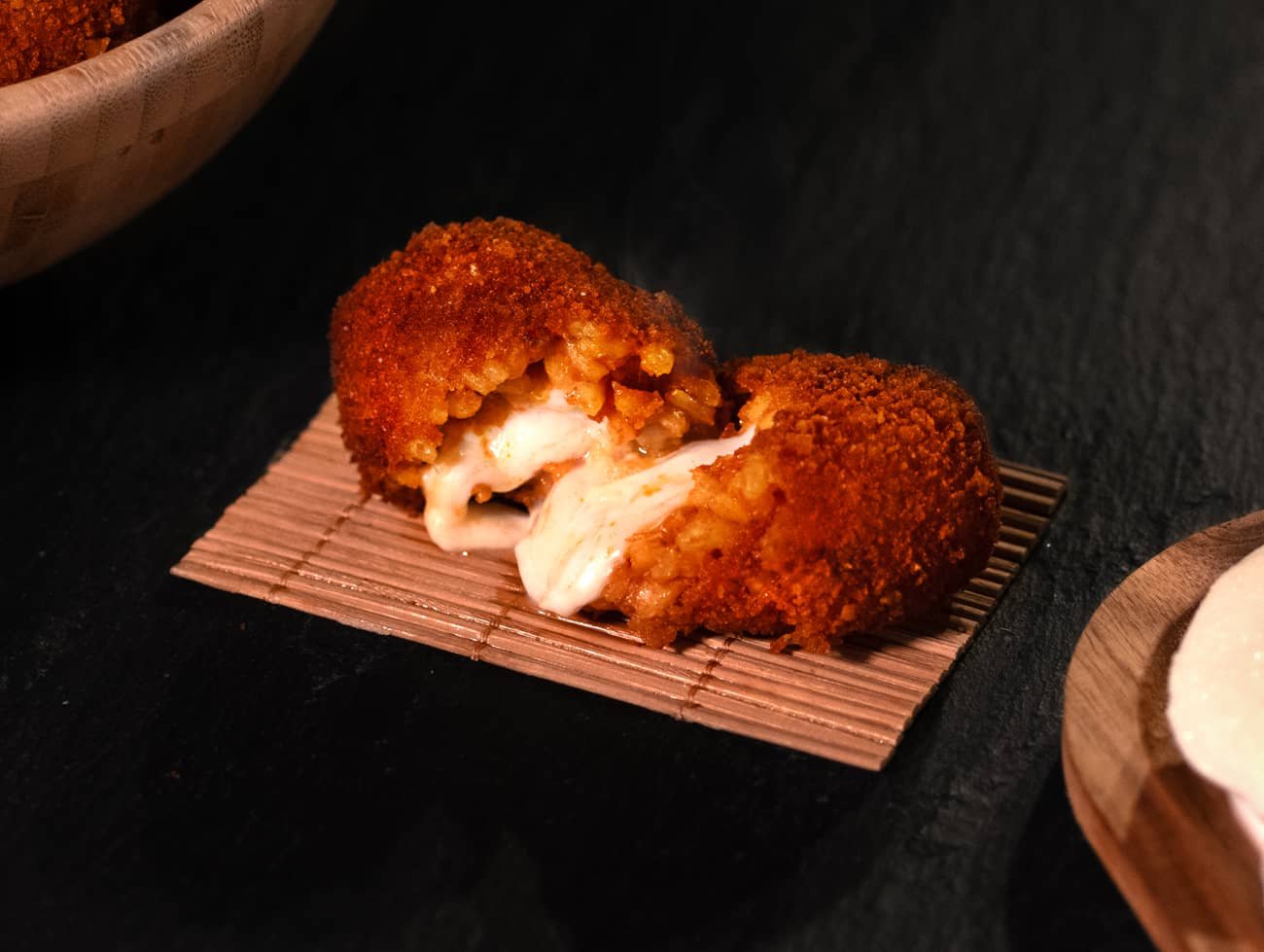 Foto del supplì con carne con vista dall'alto poggiato su una stuoietta a sua volta poggiata su un tavolo in ardesia. Il supplì è aperto a metà ed esce la mozzarella fusa.